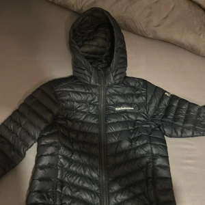 Svart dunjacka Peak Performance - Svart dunjacka från Peak Performance med huva och dragkedja framtill. Jackan har quiltad design och logga på bröstet. Perfekt för kyliga dagar och har en sportig vibe.