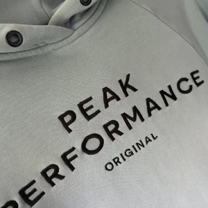 Ljusblå hoodie Peak Performance - Ljusblå hoodie från Peak Performance med svart broderad logga på bröstet. Klassisk känguruficka framtill, ribbade muddar och huva med snörningshål dock utan snören. Mjuk och skön bomullsblandning. Lite slitage vid muddarna, inget som syns när den används. 