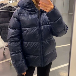 Mörkblå glansig pufferjacka/vinterjacka - Säljer en mörkblå pufferjacka med huva och dragkedja framtill. Jackan har en oversized passform och är tillverkad i glansigt syntetmaterial. Perfekt för kalla dagar och riktigt mysig att bära. Stora, fluffiga sektioner ger en cool look. Passar S/M 