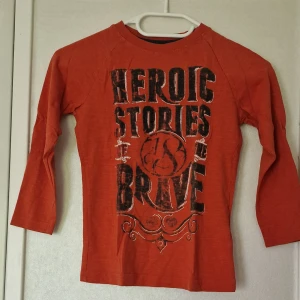 Orange långärmad tröja från Mywear Kids - Orange långärmad tröja från Mywear Kids med svart tryck framtill där det står 'Heroic Stories Be Brave'. Tröjan har rund halsringning och är tillverkad i mjuk bomull, perfekt för skolan eller chill dagar.