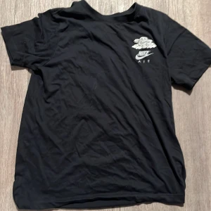 Svart Nike Air t-shirt med tryck - Svart t-shirt från Nike Air med coolt tryck på ryggen av en sneaker som skjuter upp som en raket och måne i bakgrunden. Framtill finns ett mindre tryck med moln och Nike Air-logga. Klassisk rund hals och korta.