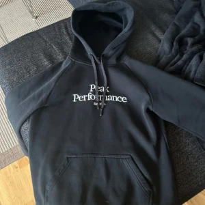 Svart hoodie från Peak Performance - Svart hoodie från Peak Performance med vit broderad logga på bröstet. Klassisk modell med huva, snörning och stor magficka framtill. Perfekt för chill dagar eller när du vill ha en enkel och snygg look. Storlek S