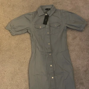 Blå kort jeansklänning Vero Moda - Snygg blå kortklänning från Vero Moda i jeansmaterial. Klänningen har puffiga korta ärmar, krage och knappar hela vägen framtill. Två bröstfickor med lock och markerade sömmar ger en cool och trendig look.