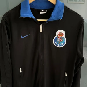Nike FC Porto svart ziptröja - Svart ziptröja från Nike med blå krage och FC Porto-emblem på bröstet. Tröjan har dragkedja framtill, två fickor med dragkedja och en framtill. Liten defekt som du ser på andra bilden!