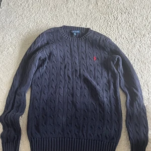 Mörkblå kabelstickad tröja Ralph Lauren - Kabelstickad mörkblå tröja från Ralph Lauren med klassisk rund hals och röd broderad logga på bröstet. Tröjan har långa ärmar och breda ribbade muddar vid ärmslut och nederkant. Perfekt för lager-på-lager under höst och vinter och den är äkta. 