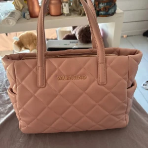 Quiltad rosa handväska från Mario Valentino - Superfin quiltad handväska i ljusrosa från Mario Valentino. Väskan har två handtag, dragkedja upptill och snyggt rutmönster. Tillverkad i mjukt syntetmaterial och har praktiska sidofickor, 3 fack på insidan och ett långt handtag medkommer. Perfekt för dig som vill ha en trendig och rymlig väska.