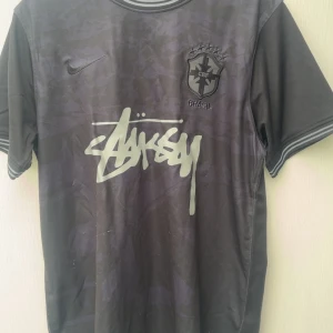 Svart Brasil x Stüssy x Nike fotbollströja - Unik svart Brasil-fotbollströja från Nike och Stüssy med grå Stüssy-logga på bröstet och Brasil-märke. Kortärmad modell med subtilt camouflagemönster och randiga detaljer vid krage och ärmslut. Perfekt för dig som gillar streetwear och fotboll.