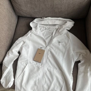 Vit Nike Dri-Fit vindjacka med huva - Snygg vit vindjacka från Nike med Dri-Fit-teknologi, perfekt för träning. Jackan har huva, hel dragkedja framtill och diskret Nike-logga på bröstet. Materialet är lätt och andas, vilket gör den grym för aktiva dagar. Stilren design och sportig vibe.