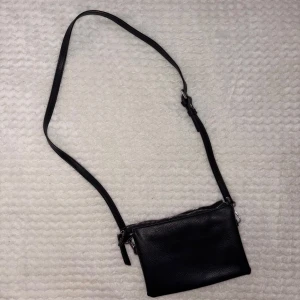 Svart axelväska i skinn med dragkedja - En stilren svart axelväska i äkta skinn med justerbart axelband och silverfärgade detaljer. Väskan har en enkel, rektangulär form och stängs med dragkedja upptill. Perfekt storlek för det viktigaste och passar till många olika outfits.