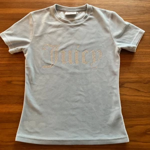 Ljusblå Juicy Couture t-shirt med strass - Ljusblå t-shirt från Juicy Couture i mjuk velour med korta ärmar. Framsidan har texten 'Juicy' i glittrande strass. Klassisk rund halsringning och figurnära passform. Perfekt för dig som vill ha en ikonisk och trendig look.
