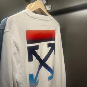 Vit sweatshirt från Off-White med tryck - Vit sweatshirt från Off-White med ikoniskt stort grafiskt tryck i rött och blått på ryggen och liten blå logga framtill. Tröjan har rund hals, långa ärmar och är gjord i mjuk bomull. Perfekt för dig som gillar streetwear och vill sticka ut. Nästan aldrig använd. Hör av er om ni har några frågor!
