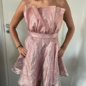 Rosa glittrig axelbandslös klänning - Supersnygg axelbandslös kort klänning i glittrigt rosa tyg med markerad midja och voluminös kjol. Klänningen har en unik struktur upptill med veckade detaljer som ger en cool och modern look. Perfekt för dig som vill sticka ut!