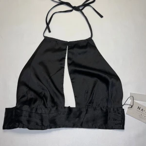 Svart halterneck topp från NA-KD - Säljer en svart halterneck topp från NA-KD i glansigt satinliknande material. Toppen har öppen rygg, knytband i nacken och bred knytning i midjan. Snygg detalj med djup slits framtill som ger en cool look.