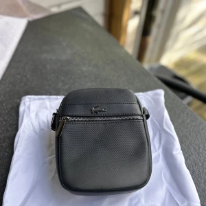 Svart axelväska från Lacoste - Snygg svart axelväska från Lacoste med klassisk krokodillogga i metall framtill. Väskan har en stilren, fyrkantig form, justerbar axelrem och praktisk dragkedja. Tillverkad i slitstark polyester med detaljer i skinnimitation. Perfekt för att bära det viktigaste med stil.