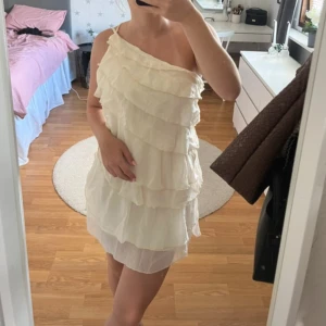 Vit volangklänning oneshoulder - Supersöt kort vit klänning med volanger och oneshoulder-design. Klänningen har tunna axelband och är tillverkad i ett lätt, luftigt material som ger en romantisk känsla. 