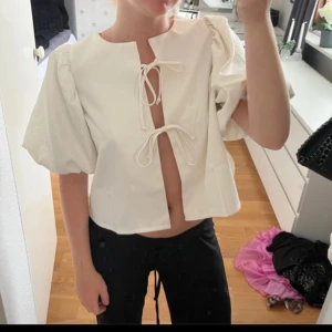Vit blus med puffärm och knyt - Säljer en vit blus med korta puffärmar och två knyt framtill. Blusen har en luftig passform med snygga detaljer som ger en trendig look. Perfekt för dig som gillar stilrena och unika toppar.