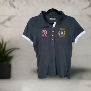 Mörkgrå pikétröja från U.S. Polo Assn. - Mörkgrå pikétröja från U.S. Polo Assn. med broderad logga och siffra 3 på bröstet. Klassisk krage, knappar framtill och vita detaljer vid ärmslut. Lite urtvättad