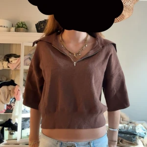 Brun stickad tröja med half zip - Snygg brun stickad tröja med halv dragkedja och krage. Croppad modell med korta ärmar, perfekt för dig som gillar en chill och trendig stil. Passar till jeans eller kjol och har en mjuk känsla mot huden.