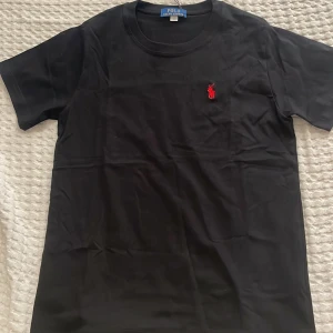 Svart t-shirt från Polo Ralph Lauren - Svart t-shirt från Polo Ralph Lauren med klassisk rund hals och korta ärmar. På bröstet finns den ikoniska röda Polo-loggan broderad. T-shirten är gjord i mjuk bomull och har en clean, stilren look som funkar till allt.