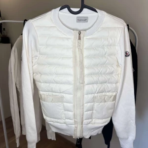 Moncler Cardigan - Snygg vit cardigan jacka från Moncler med quiltad framsida och stickade ärmar. Jackan har dragkedja framtill, rund hals och två fickor med knappstängning. Moncler-logga på ärmen ger extra stilpoäng. Perfekt för dig som gillar clean och exklusiv look.