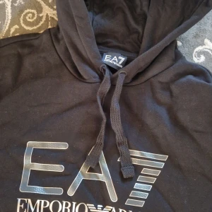 Svart EA7 hoodie från Emporio Armani - Svart hoodie från Emporio Armani EA7 med stor logga framtill. Tröjan har huva med snörning och är tillverkad i mjuk bomull. Perfekt för dig som gillar stilrena och klassiska streetwear-plagg med exklusiv känsla. Storleken är XL men jag skulle säga att passformen är en large-stor medium. Pris kan diskuteras.