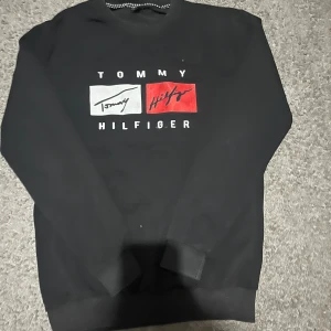 Svart Tommy Hilfiger sweatshirt S - Svart sweatshirt från Tommy Hilfiger i storlek S. Tröjan har rund halsringning, långa ärmar och en stor logga framtill i vitt och rött. Tillverkad i mjuk bomullsmix, perfekt för en chill och stilren look.