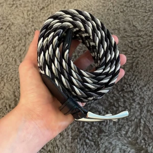 Svart och vit flätad bälte med silverdetaljer - Snyggt flätat bälte i svart och vitt med silvrigt spänne. Bältet har detaljer i svart skinn vid spännet och en cool, tvinnad design som ger en modern känsla. Perfekt för att lyfta vilken outfit som helst med en stilren touch.