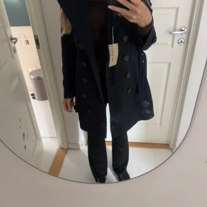 Mörkblå trenchcoat  - Säljer en klassisk mörkblå trenchcoat  med det ikoniska rutiga fodret. Jackan har dubbelknäppning, bälte i midjan och vid ärmslut samt stora slag. Perfekt för dig som vill ha en tidlös och stilren jacka.