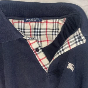 Mörkblå pikétröja från Burberry - Snygg mörkblå pikétröja från Burberry med klassiskt rutigt tyg i kragen och knappslån. Tröjan har broderad logga på bröstet och detaljer i vitt och rött. Perfekt för dig som gillar stilrena och tidlösa plagg.