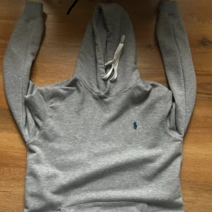 Grå hoodie från Ralph Lauren - Säljer en klassisk grå hoodie från Ralph Lauren med blå logga på bröstet. Tröjan har huva med vita snören. Storlek Xs