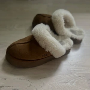 Bruna UGG tofflor med fårull - Mysiga bruna UGG tofflor med fluffig vit fårullskant. Gjorda i mjuk mocka och har en platt sula. Perfekta för att hålla fötterna varma under kyliga dagar. Klassisk UGG-logga på sidan och rund tå för extra komfort.