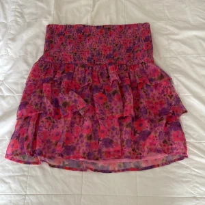 Blommig volangkjol från Bikbok - Säljer en kort kjol från Bikbok med blommigt mönster i rosa, lila och röda toner. Kjolen har bred smock i midjan och flera lager med volanger som ger en cool och lekfull look. Perfekt för dig som gillar färg och mönster.