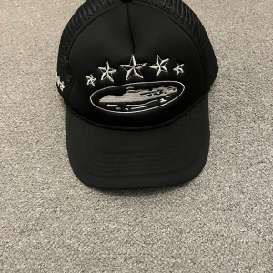 Svart truckerkeps från Corteiz - Svart truckerkeps från Corteiz med broderade vita stjärnor och logga framtill. Kepsen har mesh på sidorna och baktill för extra ventilation samt justerbar snapback. Broderad signatur bak och detaljerade brodyrer på sidorna. Perfekt för en streetwear-look.