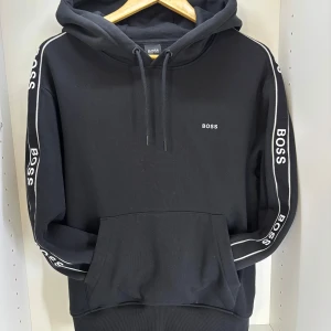 Svart hoodie från BOSS - Svart hoodie från BOSS med vit logga på bröstet och längs båda ärmarna. Tröjan har huva med snörning och en stor magficka. Klassisk och stilren design med ribbade muddar och bekväm passform. Knappt använd, säljs eftersom den inte passar mig.