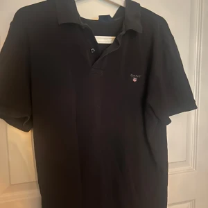 Svart pikétröja från Gant - Klassisk svart pikétröja från Gant med korta ärmar och liten broderad logga på bröstet. Tröjan har krage och knäppning framtill. Perfekt för en clean och stilren look.