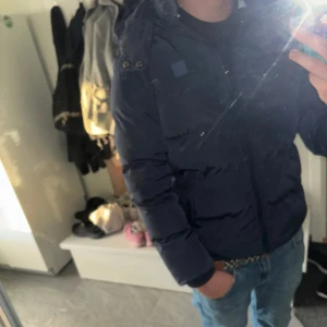 Mörkblå pufferjacka från Urban classics - Säljer en mörkblå pufferjacka från urban classics med huva och dragkedja framtill. Jackan är vadderad och har en clean look med diskreta detaljer. Perfekt för kalla dagar och riktigt skön att bära. Snygg passform och enkel att matcha med jeans eller hoodies.