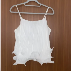 Vit plisserad linnetopp från Vero Moda - Supersöt vit linnetopp från Vero Moda med tunna axelband och plisserat tyg. Toppen har vågiga volangdetaljer längst ner som ger en cool och lekfull look. Perfekt för varma dagar när du vill sticka ut lite extra.