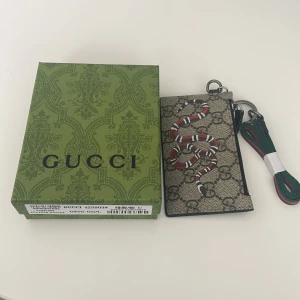 Gucci plånbok med orm och GG-mönster - Snygg plånbok från Gucci med klassiskt GG-mönster och en cool röd, vit och svart orm på framsidan. Baksidan är svart med flera kortfack. Plånboken har en metallring och medföljande grön-röd rem. Perfekt för dig som vill ha något unikt och lyxigt.