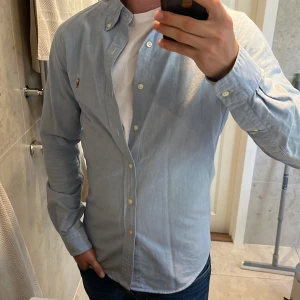 Ralph Lauren Skjorta - Säljer en stilren ljusblå skjorta från Ralph Lauren i slim fit-modell. Skjortan har klassisk button down-krage, broderad logga på bröstet och knäppning framtill. Tillverkad i mjuk bomull och har långa ärmar med manschettknappar.