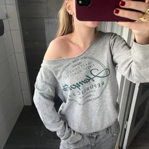 Grå offshoulder tröja Hampton Republic - Grå sweatshirt från Hampton Republic med tryck framtill. Tröjan har en snygg offshoulder-modell och långa ärmar. Mjuk och skön i bomullsblandning, perfekt för chill dagar. Bred mudd vid ärmslut och nederkant för en avslappnad look.