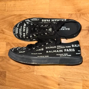 Balmain skor - Svarta sneakers från Balmain Paris med vit logotyptext tryckt över hela skon. Klassisk låg modell med snörning och platt sula. Ovandel i textil och sula i gummi. Snygga och trendiga skor som sticker ut med sin tydliga branding.