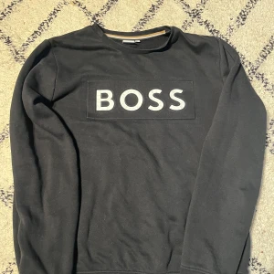 Svart sweatshirt från BOSS med tryck - Svart sweatshirt från BOSS med stort vitt BOSS-tryck på bröstet. Tröjan har rund halsringning, långa ärmar och en klassisk passform. Perfekt för en stilren och avslappnad look.