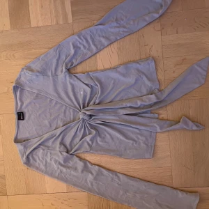 Ljusgrå omlottopp med knyt - Säljer en ljusgrå omlottopp från Gina Trico med långa ärmar och knyt framtill. Toppen har en djup v-ringning och är tillverkad i mjuk bomullsmix. Perfekt att styla med jeans eller kjol för en chill look. Har dock ett litet hål på baksidan