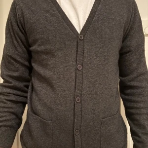 Mörkgrå kofta från Lawrence Grey - Mörkgrå kofta från Lawrence Grey med v-ringning, knäppning framtill och två fickor. Klassisk och stilren modell i mjukt material, perfekt för lager-på-lager. Långärmad och ribbade muddar vid ärmslut och nederkant.