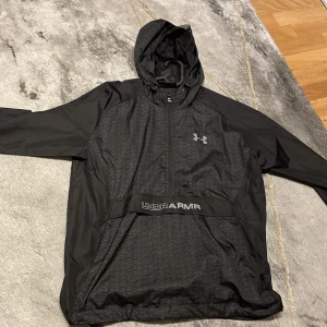 Svart träningsjacka med huva från Under Armour - Säljer en svart träningsjacka från Under Armour med huva och dragkedja. Jackan har logga på bröstet och textdetalj på magen. Perfekt för träning och har en sportig look med lång ärm och lätt material. Pris kan diskuteras 😊