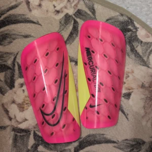Nike Mercurial Lite benskydd med strumpor och påse - Säljer ett par Nike Mercurial Lite benskydd i stark rosa med svart och gul detalj, samt tillhörande svarta strumpor med neongult Nike swoosh. Benskydden har ett hexagonmönster och är tillverkade i lätt plast med mjuk insida. Kommer med original svart förvaringspåse.