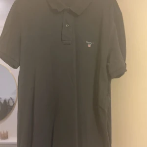 Svart pikétröja från GANT - Svart pikétröja från GANT med klassisk krage och två knappar framtill. Tröjan har korta ärmar och GANT-logga med sköld på bröstet. Tillverkad i mjuk bomull som är skön mot huden. Perfekt för en clean och stilren look.