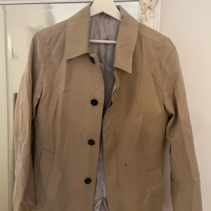 Beige rock med rutigt foder - Stilren beige rock med klassisk krage och svarta knappar. Jackan har ett rutigt innerfoder i grå toner och två sidfickor. Perfekt för dig som gillar en clean och tidlös look. Obs finns en fläck på jackan, har inte testat att tvätta den så borde förhoppningsvis gå bort! 