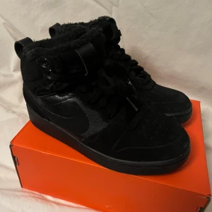 Svarta Nike high top sneakers med fuskpäls - Svarta Nike high top sneakers med fluffigt fuskpälsfoder, snörning och robust platt sula. Snyggt ton-i-ton med olika material som mocka och syntet, samt klassisk Nike-logga på sidan. Perfekt för dig som vill ha streetstyle med extra värme.