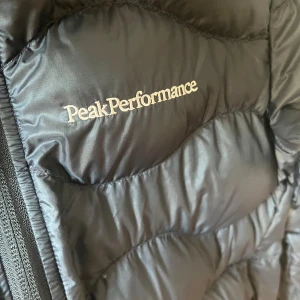 Svart dunjacka Peak Performance - Svart dunjacka från Peak Performance med quiltad design och dragkedja framtill. Jackan har hög krage och diskret logga broderad på bröstet. Perfekt för kalla dagar och har en stilren look som funkar till det mesta.